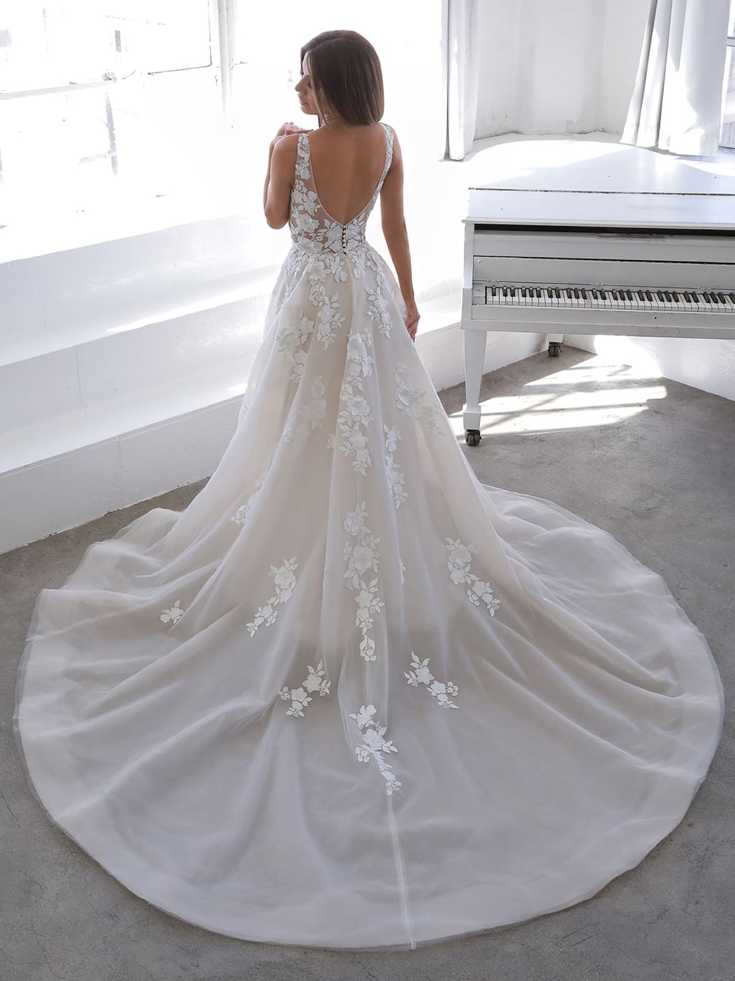 Web Nala, an ivory and nude A-line wedding gown by Blue by Enzoani, featuring a plunging V-neckline, low V-back, and delicate floral 3D appliqués meticulously scattered across the bodice and flowing organza skirt, which extends into a cathedral train.
