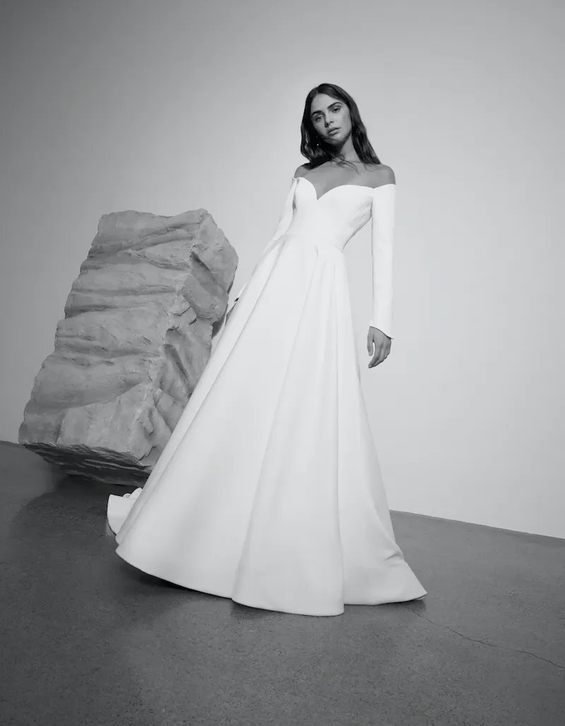 Leah, an ivory bridal gown by Alyne, features a romantic off-the-shoulder sweetheart neckline with long sleeves. The elegant crepe fabric forms an A-line ball gown skirt with a cathedral train. The dress is modeled in a black and white studio image, with the model posing confidently next to a large textured rock sculpture.