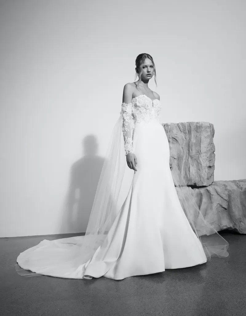 Vivian, an ivory mermaid bridal gown with an off-the-shoulder sweetheart corset bodice featuring intricate lace applique on the 3/4 length sleeves, and a flowing satin and crepe sheath skirt creating a dramatic cathedral-length train. Designed by Alyne.