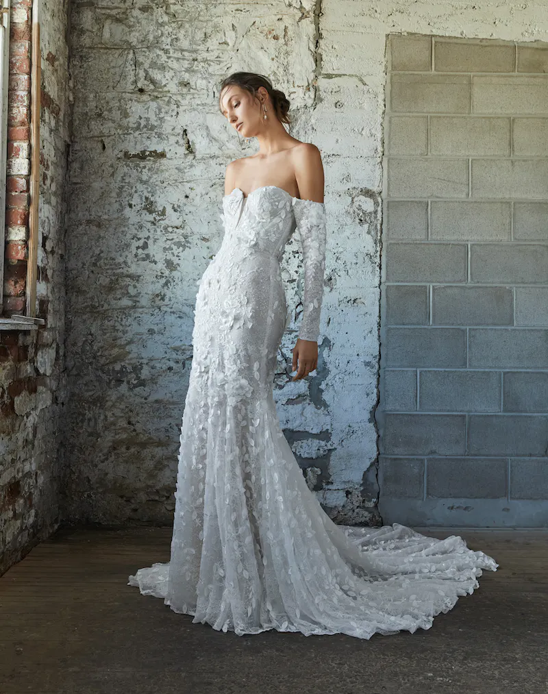 Lilibet, an ivory and nude fit-and-flare bridal gown by Rivini, features a strapless sweetheart bodice with detached long off-the-shoulder illusion sleeves. The dress is intricately covered in delicate lace appliques and subtle beading, creating a textured pattern that extends down to its chapel-length train. The model stands elegantly posed in a rustic setting with exposed brick and concrete walls.
