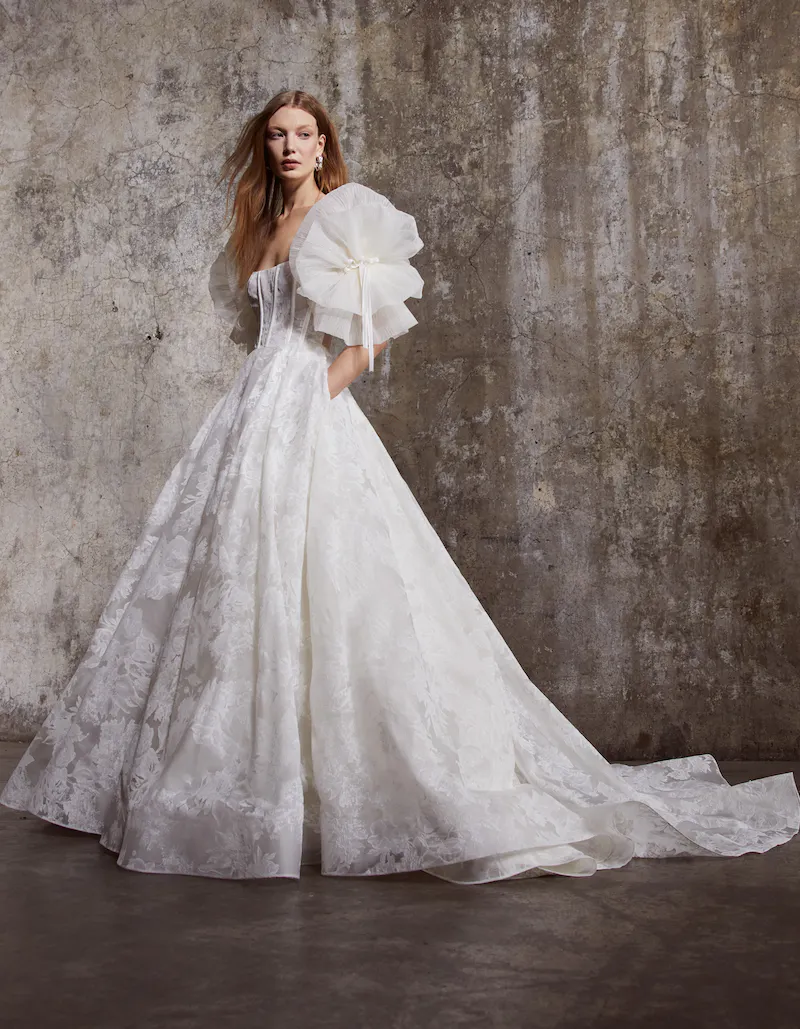 1669174099433-Colette Colette, an ivory Rivini bridal gown. This dramatic ball gown features a strapless, modified sweetheart corset bodice and voluminous, ruffled organza off-the-shoulder sleeves with pearl and ribbon embellishments. The skirt, crafted from embroidered silk and organza, extends into a chapel-length train. The model stands with one hand in a pocket.