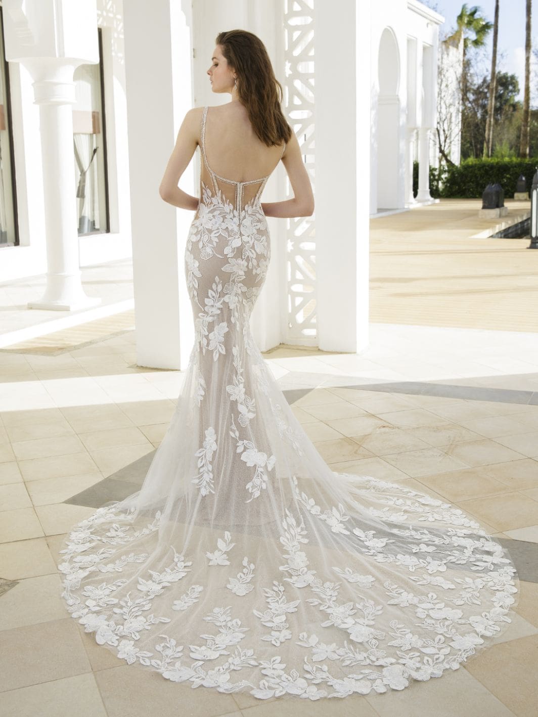 1686847976500-f157c40b-ede4-4f08-b97b-5bde0dceb136 Rosemary, an Ivory/Nude/Nude sheath wedding gown that shines with dazzling details. It features a square neckline, supportive beaded spaghetti straps, and a breathtaking low, corset-style back adorned with exquisite beaded trim. Striking embroidered lace is strategically placed over the sheer bodice and skirt, complemented by illusion side cut-outs designed to enhance curves. The gown flows into a chapel-length train embellished with delicate lace appliques. The model is seen from the back, looking over her left shoulder, in a bright outdoor architectural setting.