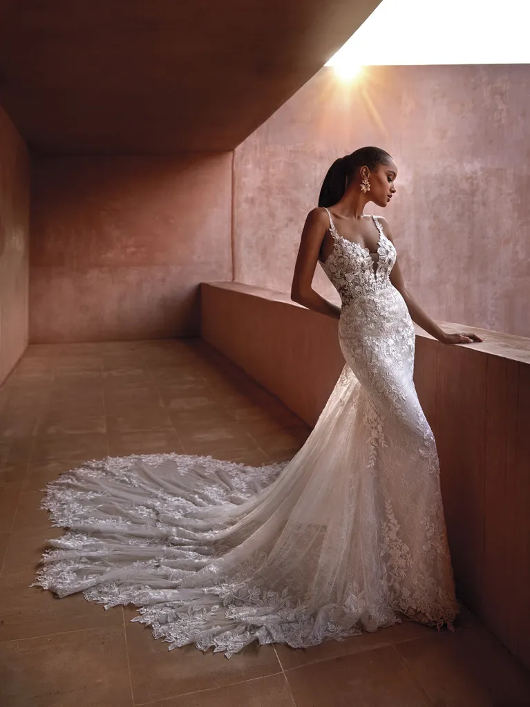 SEYCHELLES, an Off White/Nude Pronovias bridal gown with a mermaid fit-n-flare silhouette, featuring a plunging V-neck, spaghetti straps, and a low back. The dress is richly embellished with intricate lace and beading, extending into a magnificent cathedral-length train. A model with dark skin and a high ponytail is wearing the gown, posing gracefully in a warm, sunlit, terracotta-hued architectural space.