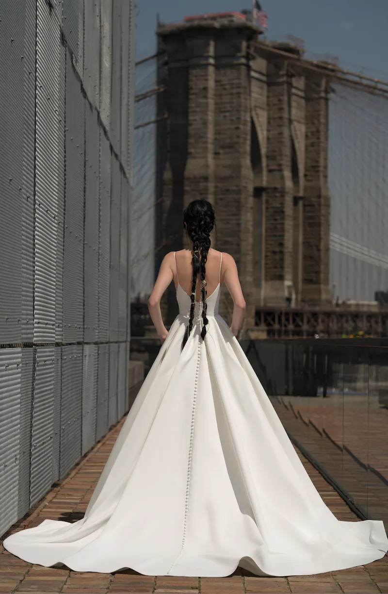 1698349910532-b66b80f7-0f7c-46c3-8a1a-34484ec022fb_91916_CRAWFORD_B Crawford, an ivory matte satin bridal gown with slim spaghetti straps, a low back, and a long line of buttons extending down its voluminous ballgown skirt, worn by a woman with long braided hair, standing with her back to the camera and looking towards the Brooklyn Bridge.