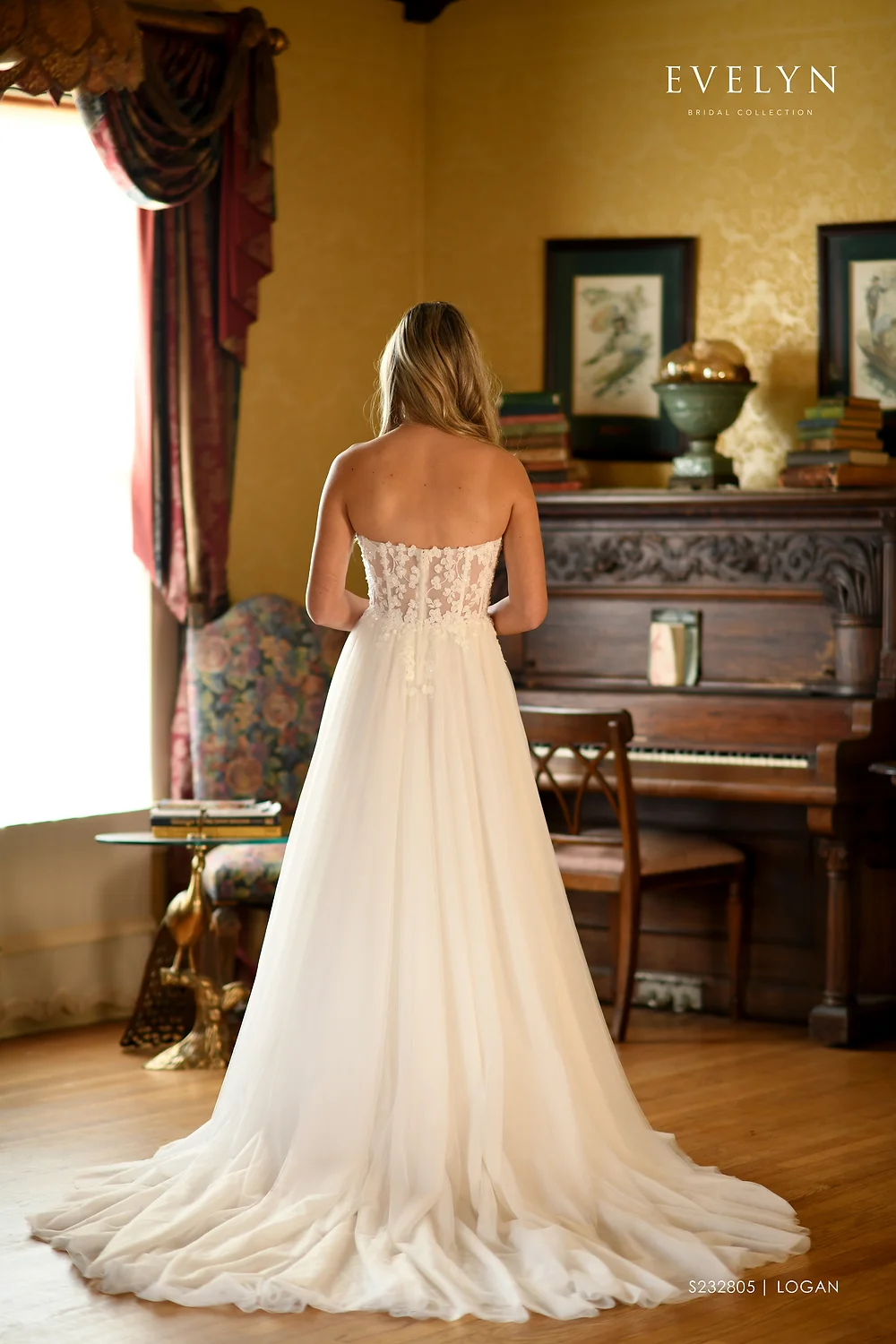 1698422107190-0baeb4-a23c9ff61cc2412e9962fc8c59e6e09a JoJo, an Ivory/Ivory A-line wedding gown, featuring a strapless sweetheart neckline and an illusion lace bodice that transitions into a whimsical soft tulle skirt with a chapel train. The dress is modeled from the back by a woman with long blonde hair, standing in a richly decorated room with a bright window and red curtains to the left, and a grand piano and antique furnishings to the right.