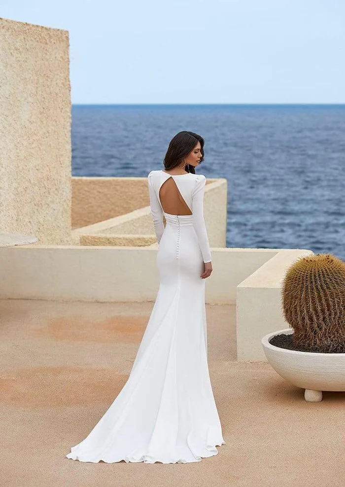 Adrienne, an off white/crystal crepe bridal gown with long sleeves, a keyhole back, and a fit and flare silhouette, featuring a line of delicate buttons down the back and a flowing train. A woman with dark hair wears the gown, standing on a light-colored terrace overlooking the blue ocean, seen from the back as she looks over her shoulder. A large potted cactus is on the right.