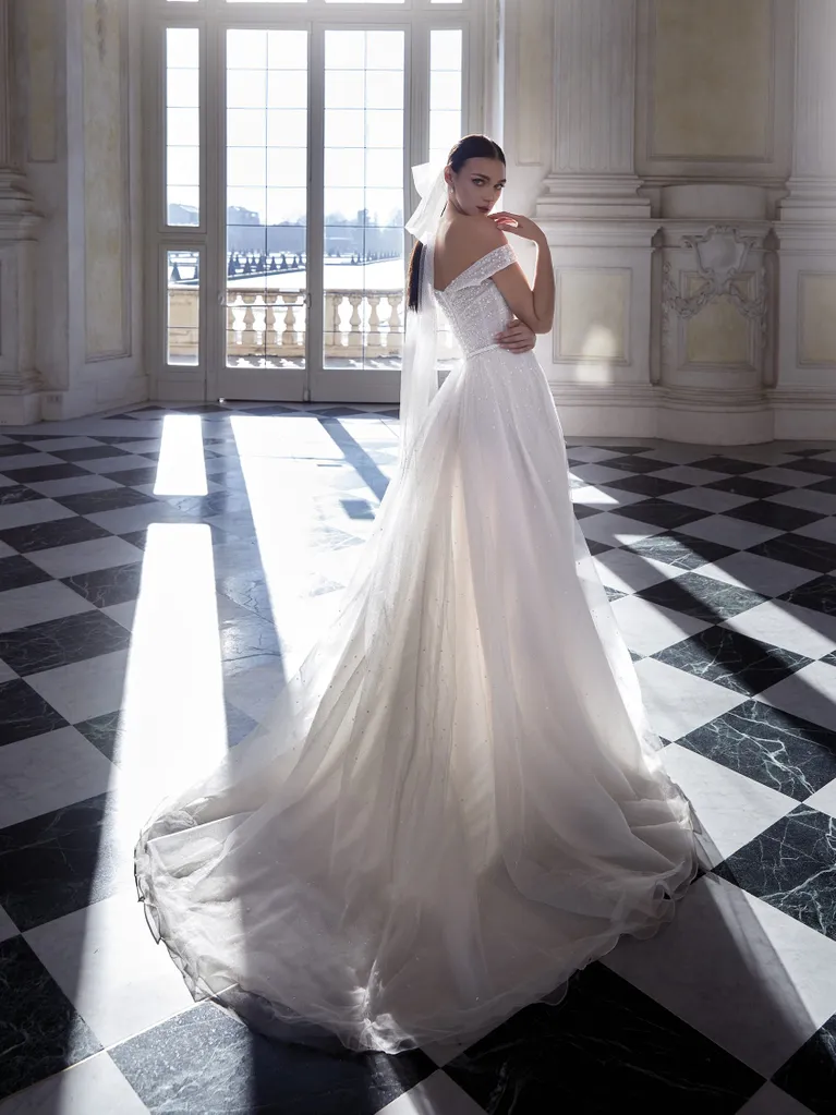 1698431453602-ida_c Ida, an OFF WHITE/LIGHT BEIGE Pronovias bridal gown, a show-stopper for the bold and glamorous bride. This dazzling gown features an off-the-shoulder neckline and is completely covered in sparkling embellishments from the bodice to the voluminous, flowing tulle skirt that extends into a cathedral train. The model is depicted from behind, looking over her shoulder, in a sunlit grand hall with a checkered floor.
