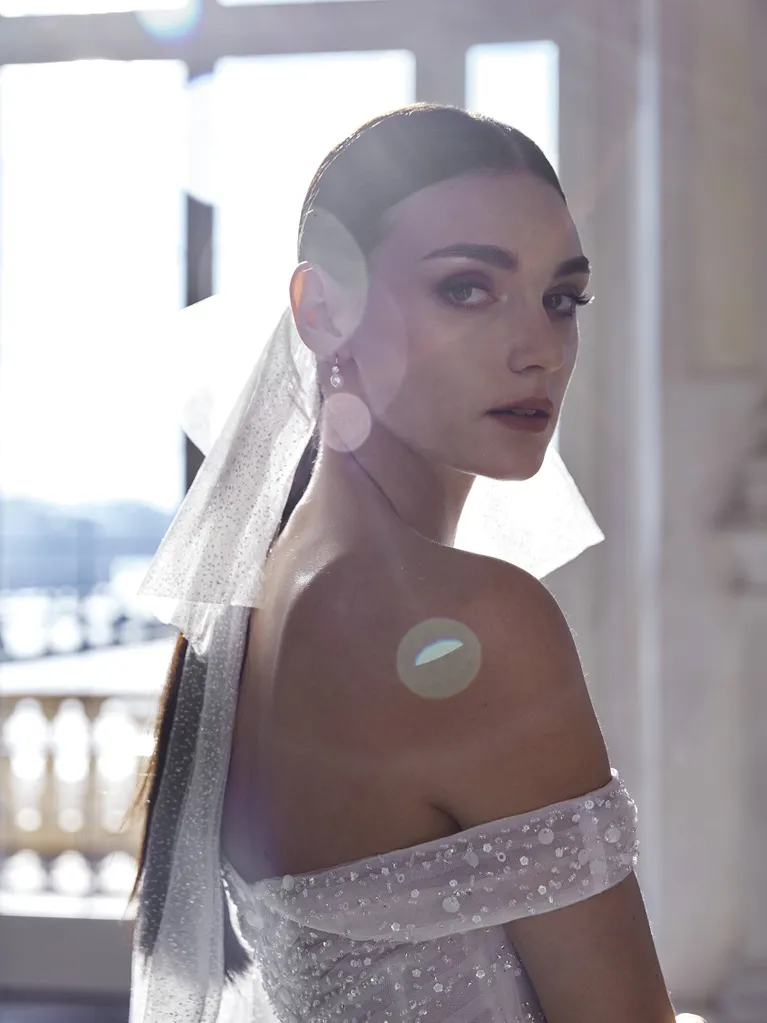1698431453603-ida_da Ida, an off-white off-the-shoulder bridal gown by Pronovias, completely covered in dazzling sparkling embellishments. The model looks elegantly over her shoulder, wearing delicate pearl earrings and a sheer, glitter-dusted veil that cascades down her back.