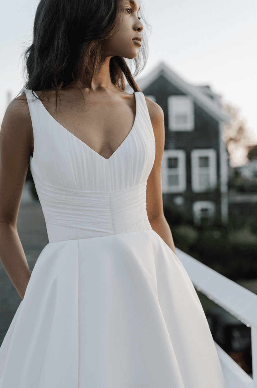 1700937064432-screen-shot-2023-11-25-at-1-28-33-pm Birdsong, an Anne Barge silk white A-line bridal gown featuring a V-neck and a finely pleated chiffon bodice above a smooth satin skirt, worn by a model with dark hair looking contemplatively to the right, bathed in warm golden light outdoors.