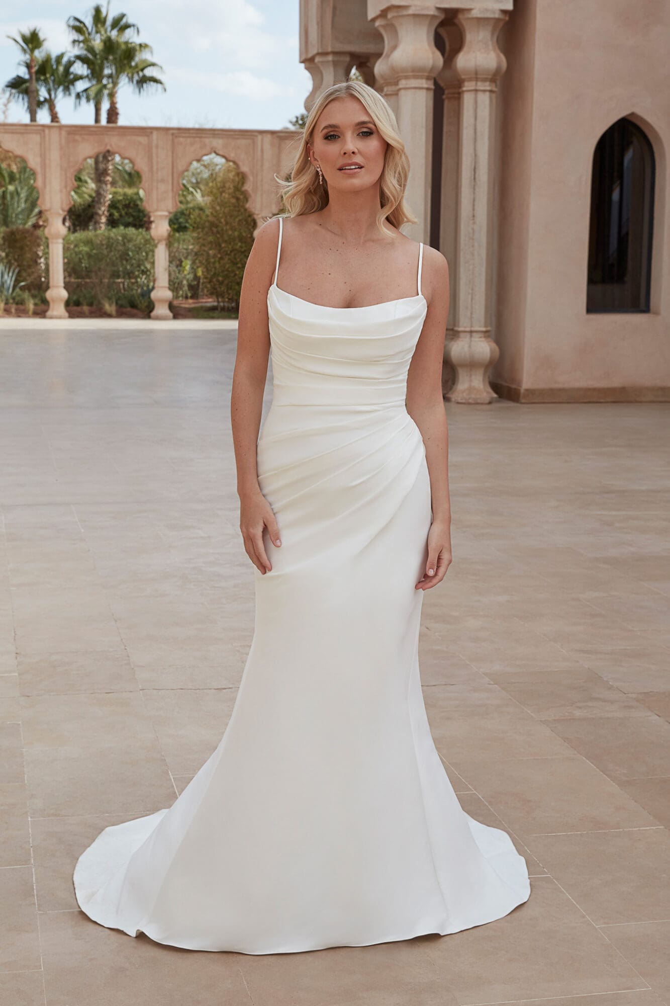 1701967912129-sincerity-bridal 44424, an ivory Mikado fit-and-flare bridal gown by Sincerity by Justin Alexander, featuring delicate spaghetti straps, a draped scoop neckline, and a chapel train, is worn by a blonde model who stands on a tiled patio, with arched stone architecture and palm trees visible in the sunny background.