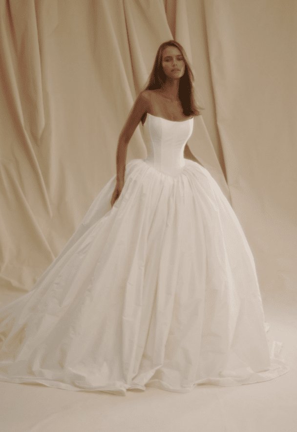 1713371588056-screen-shot-2024-04-17-at-12-32-03-pm Senstudio SE032, an ivory strapless taffeta ball gown bridal gown featuring a structured Basque bodice with corset detailing, and a voluminous, modest cathedral-length skirt, worn by a model.