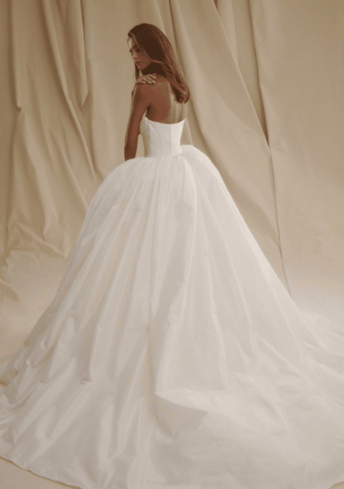 1713371588057-screen-shot-2024-04-17-at-12-32-11-pm SE032, an elegant ivory taffeta strapless ball gown by Senstudio. The dress features a structured bodice with a basque waist, a voluminous skirt with bold pleating, and a long cathedral train. A model with brown hair is shown from the back, looking over her left shoulder.