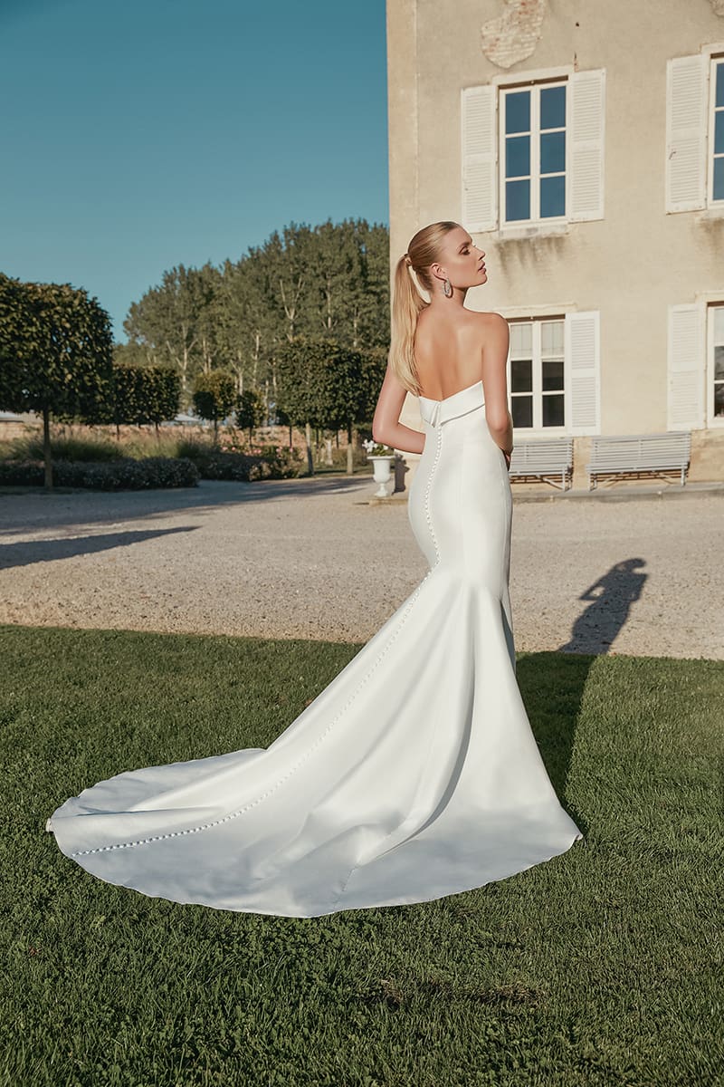 1718999534763-img-44456-fb-sincerity-bridal Marilyn, an ivory strapless fit and flare wedding gown crafted from stretch Mikado by Sincerity by Justin Alexander. The bride, with blonde hair in a ponytail, is seen from the back, showcasing the gown's elegant cuffed neckline and a long row of buttons that extend down the back and along its chapel train, which flows across green grass. She stands outdoors in a sunlit garden, with a rustic building, gravel path, and trees in the background.