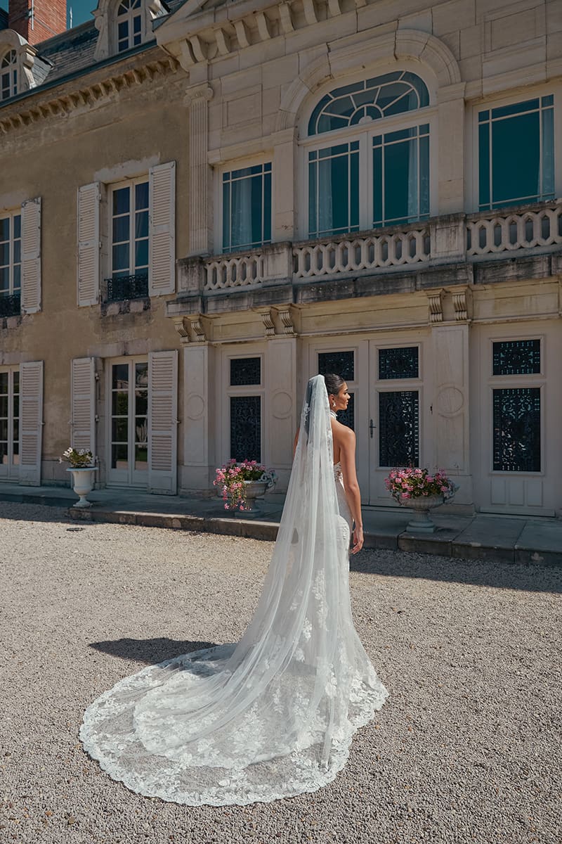 1718999721454-img-44467-tg1053-fb-sincerity-bridal Madeline, a sand, ivory, and latte fit and flare bridal gown by Sincerity by Justin Alexander, with intricate lace and shimmering sequins. The bride, with an elegant updo, is seen from behind, walking on a gravel path, showcasing the gown's elegant chapel train and a long, sheer veil with matching lace trim. The backdrop is a magnificent historic stone mansion with large arched windows, a decorative balcony, and classic doors.