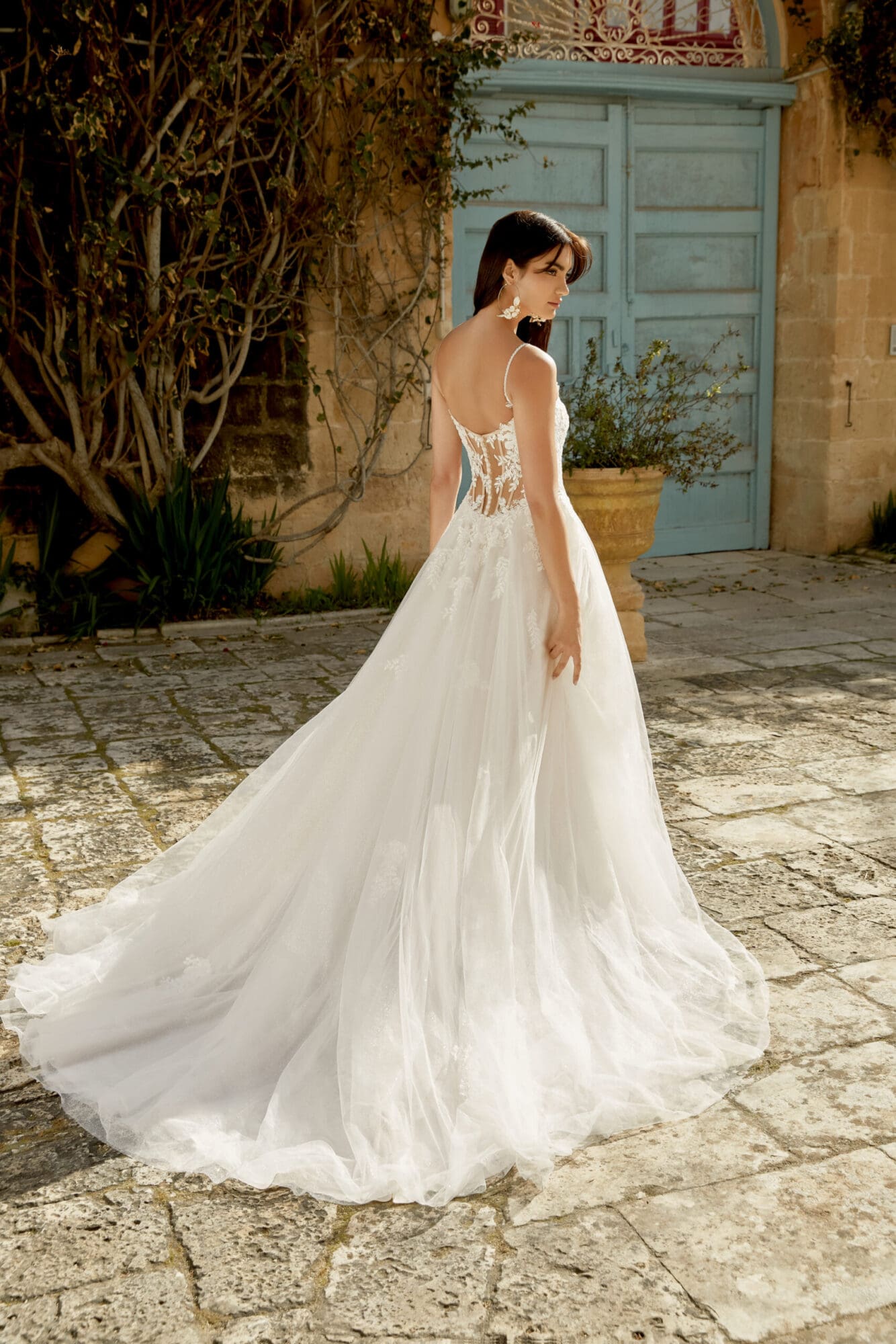 1722026756281-sin_44487_fb Maggie, a Pink Champagne, Ivory, and Latte A-line bridal gown by Sincerity by Justin Alexander, is seen from the back on a model with dark hair and floral hoop earrings. The dress features a romantic illusion lace bodice with spaghetti straps and a basque waistline, leading to a chapel-length skirt made of sparkling glitter tulle and delicate lace. She stands in a sunlit outdoor courtyard with weathered stone paving, old walls, and a light blue door.