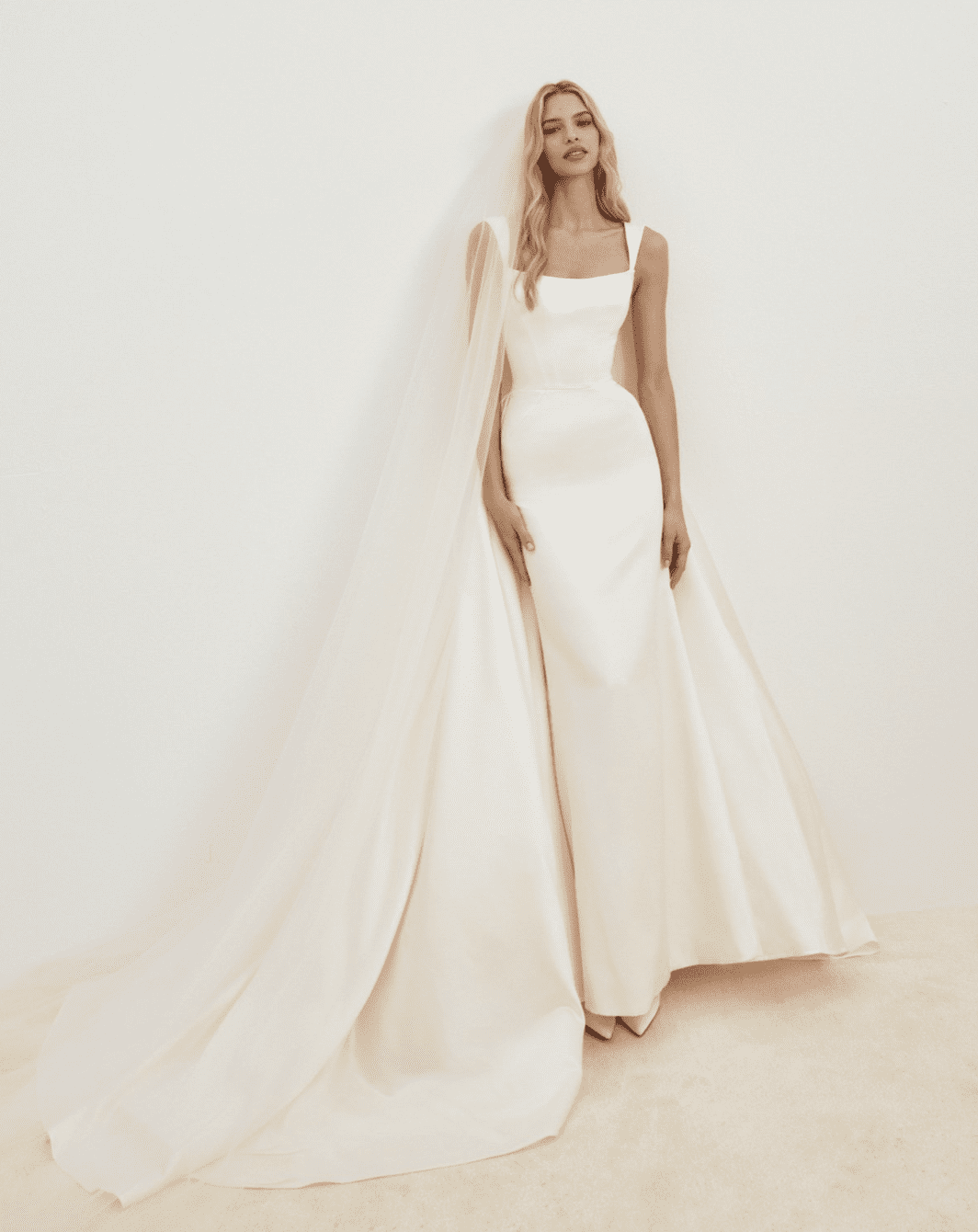 1737837880040-screen-shot-2025-01-25-at-3-43-45-pm SE042, a cream fit n flare satin wedding gown featuring a square neckline, a cathedral train, and a delicate sheer tulle cape draped elegantly over one shoulder.