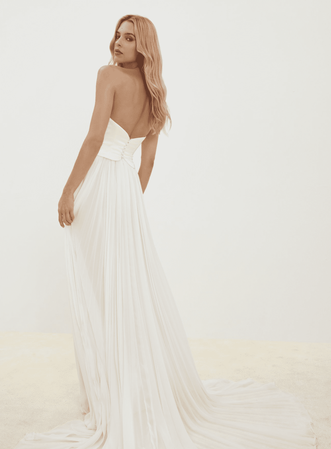 1737837982999-screen-shot-2025-01-25-at-3-45-26-pm An ivory strapless bridal gown with a structured corset bodice, featuring a row of delicate covered buttons down the back. The slim A-line silhouette flows into a pleated chiffon skirt, creating an airy and ethereal look with a chapel train, modeled by a woman looking over her shoulder.