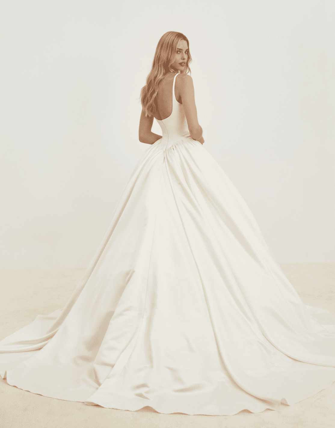 1737838170550-screen-shot-2025-01-25-at-3-49-03-pm SE045, an ivory satin bridal ballgown with a structured Basque waist, a scoop back, and delicate spaghetti straps, features a full pleated skirt flowing into a cathedral train. The model is photographed from behind, looking over her shoulder.