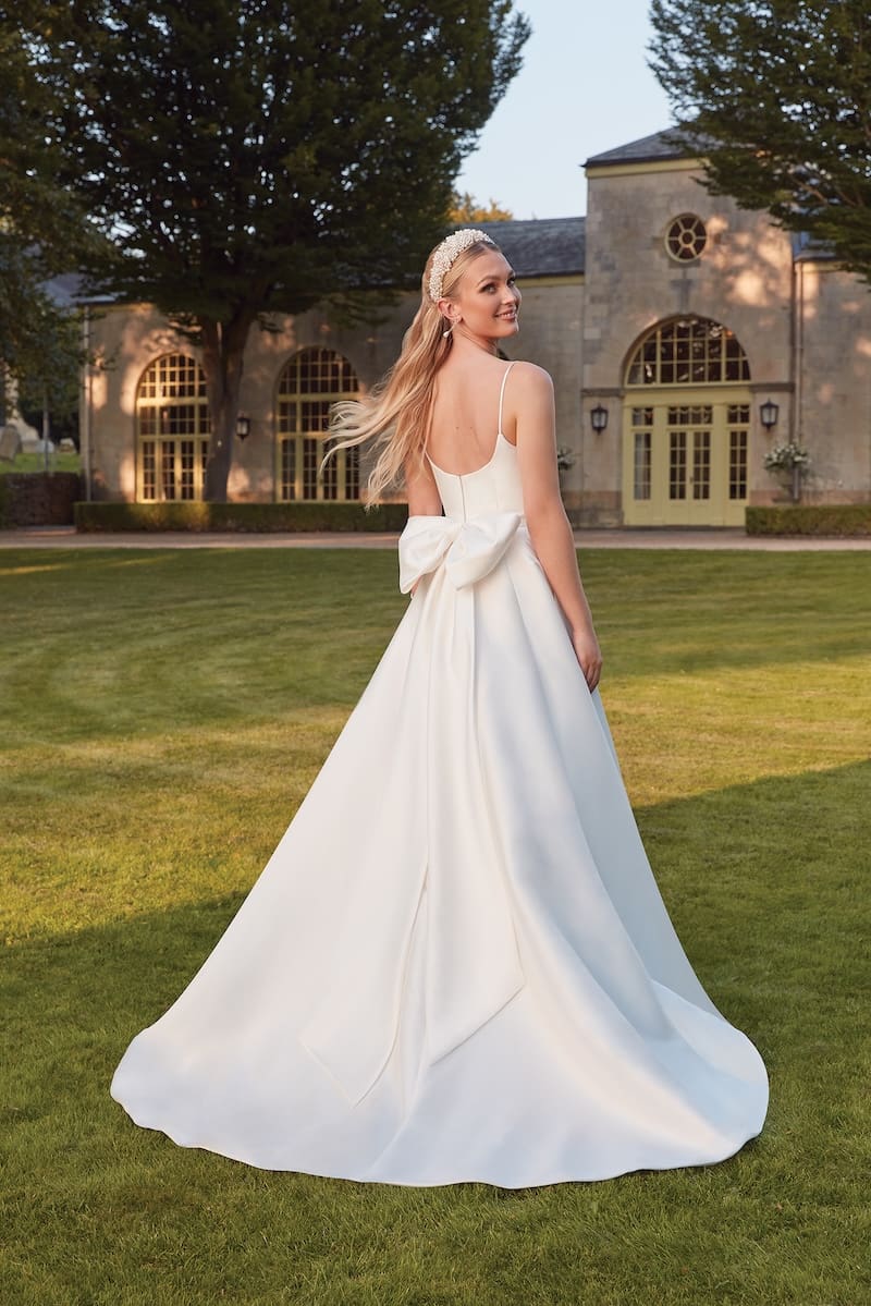 1738780078984-mongomery-sincerityja3 Montgomery, an ivory A-line bridal gown by Sincerity by Justin Alexander, crafted from Mikado. It features a square neckline, delicate spaghetti straps, a natural waist adorned with a large, elegant back bow, and a chapel train. The smiling blonde bride, wearing a pearl headband and drop earrings, looks over her shoulder, standing on a sunlit green lawn with a beautiful stone building in the background.