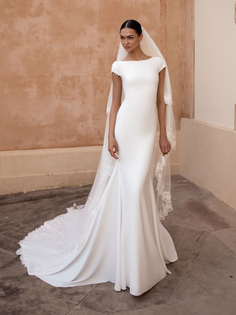 1738780287795-anitra-pronovias-front ANITRA, an off-white Pronovias bridal gown featuring a fit-and-flare silhouette, a modest bateau neckline, and delicate cap sleeves. The crepe gown showcases a chapel-length train adorned with intricate lace and embroidery, complemented by a matching lace-edged veil.