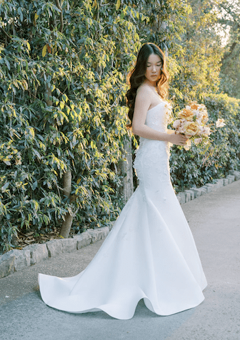 1738781541967-bella-bluewillow1 Bella, a silk white Anne Barge fit-and-flare bridal gown featuring a strapless neckline, chapel train, and delicate 3D floral organza embellishments, worn by a woman with long dark hair holding a peach and yellow floral bouquet, standing against a lush green leafy wall in warm golden light.