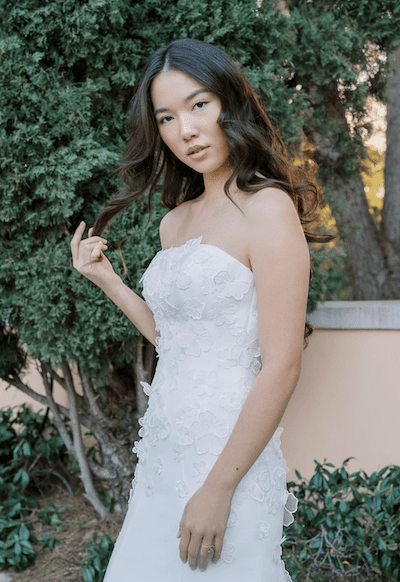 1738781541968-bella-bluewillow2 Bella, a silk white strapless fit-and-flare bridal gown by Anne Barge, featuring delicate organza 3D floral appliqués artfully scattered across its structured faille fabric, creating a soft, ethereal texture. The gown beautifully hugs the body before gracefully flaring out. A young woman with long, dark wavy hair poses elegantly in the gown, subtly touching her hair and looking directly at the camera, against a backdrop of lush green foliage and a warm-toned wall.