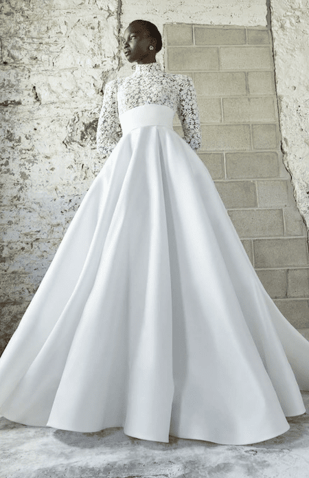 Thyra, an ivory Rivini bridal gown featuring a modest, sheer lace bodice with long sleeves and a high neckline, complemented by a wide empire waistband and a voluminous silk ball gown skirt with a chapel train. A dark-skinned model poses against a rustic white brick wall.