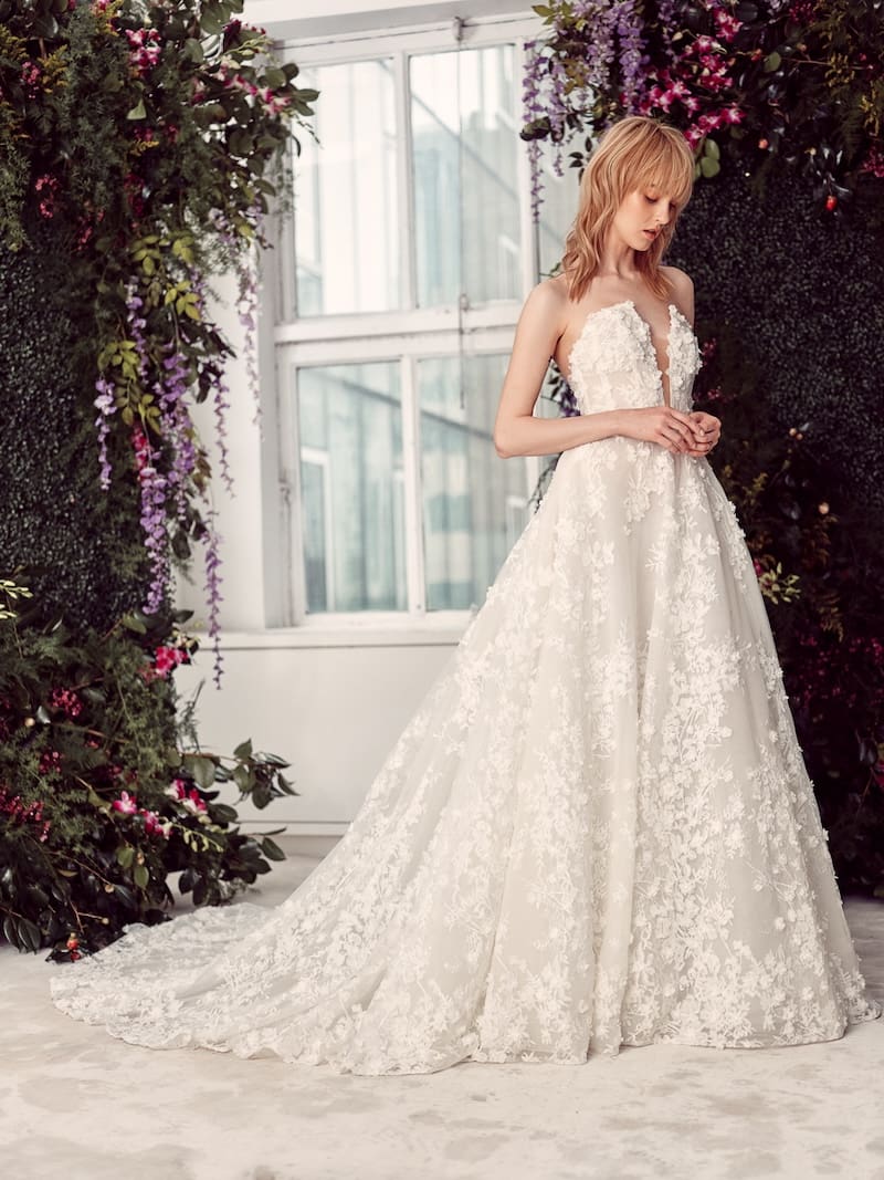 1738863104133-Reagan_Rivini Reagan, an ivory strapless ball gown, features a deep plunge neckline and is adorned with intricate 3D floral lace appliques throughout its full skirt and chapel train. The dress is modeled by a blonde woman in a lush, floral setting with a large window in the background.