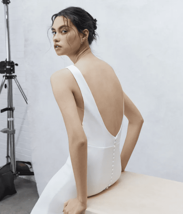 1738868023914-Luna_Alyne Luna, an ivory fit-and-flare bridal gown by Alyne, featuring a dramatic plunging back adorned with a cascading row of delicate buttons down the entire length of the train. The sleek crepe dress, designed with a timeless bateau neckline, is modeled from the back as she looks over her shoulder.