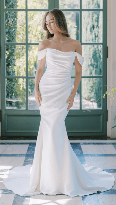 1738950241295-Naomi_AnneBarge1 Naomi, an Anne Barge bridal gown in Silk White Crepe, features a sophisticated off-the-shoulder scoop neckline with elegant draped sleeves and a ruched bodice. The fit-and-flare silhouette is form-fitting through the hips before gracefully flaring into a chapel-length train. The gown is modeled by a woman with long, flowing hair, standing on a white and blue tiled floor in front of large green-paned windows looking out onto a lush garden.