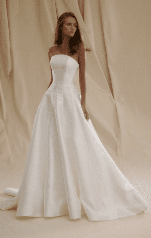 1738962362203-blair-senstudio1 Senstudio SE020, an ivory strapless A-line bridal gown crafted from Mikado fabric, featuring a natural waistline with subtle pleats and a flowing cathedral train. The gown is worn by a model with long brown hair, posing against a soft, draped beige background.