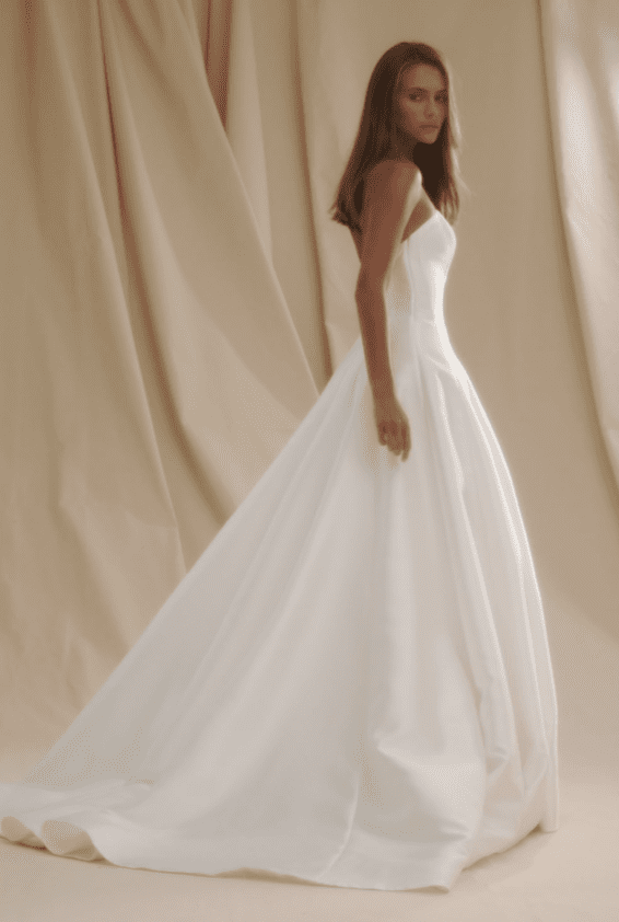 1738962362204-blair-senstudio2 SE020, an ivory strapless A-line wedding gown by Senstudio, crafted from luxurious mikado. The gown features a natural waist with decorative seams, a full skirt, and a dramatic cathedral-length train. A model with long brown hair stands facing slightly away, looking over her left shoulder, against a softly lit, draped beige background.