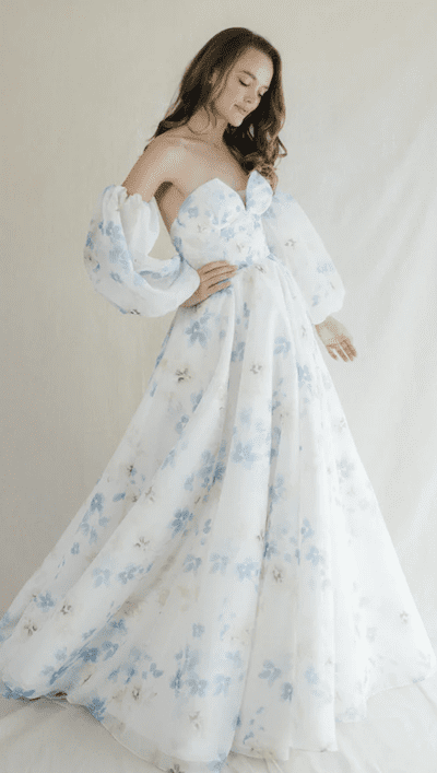 1738964945101-MorningGlory_AnneBarge-1 Morning Glory, an Anne Barge bridal ball gown crafted from watercolor printed organza. This floor-length dress features a strapless sweetheart neckline, a structured bodice, and detached voluminous 3/4 length puff sleeves. The white organza is adorned with delicate blue and subtle grey floral patterns, creating a light and airy feel.