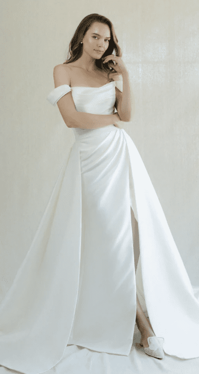 1738965009032-tucker-annebarge Tucker, a Pearl Shantung Anne Barge bridal gown, features a strapless off-the-shoulder scoop neckline with delicate ruching on the bodice and a flattering dropped waist effect. The lustrous satin design flows into a high-slit skirt, completed by a voluminous cathedral-length train, evoking a modern ball gown silhouette. The model, adorned with pearl earrings, poses elegantly with her hand to her chin.