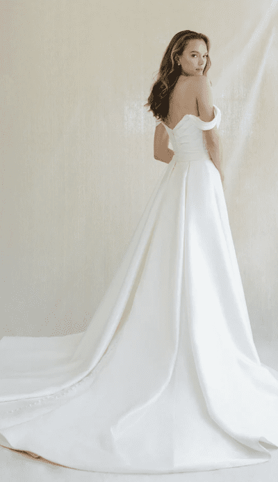 1738965134571-tucker-annebarge2 Tucker, an Anne Barge Pearl Shantung bridal gown, is a breathtaking off-the-shoulder ballgown with a dramatically draped bodice, a flattering dropped waist, and a voluminous skirt flowing into a cathedral train. The model is posed from behind, looking over her shoulder at the viewer.