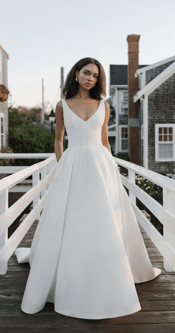1738965306790-birdsong-annebarge Birdsong, an Anne Barge silk white bridal gown featuring a V-neck pleated chiffon bodice, a voluminous Duchess Satin ballgown skirt with crisp box pleats, and a dramatic cathedral train. A model wearing the gown stands on a wooden bridge with white railings, looking at the camera, with shingled buildings in the background.
