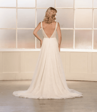 1738966976019-lady-evelyn-bridal2 Lady, an Ivory/Pearl A-line wedding gown with a deep V-neck back, featuring a bodice sprinkled with delicate pearls and a flowing, sparkling tulle skirt with a cathedral train. The model is shown from behind, looking towards a large frosted window.