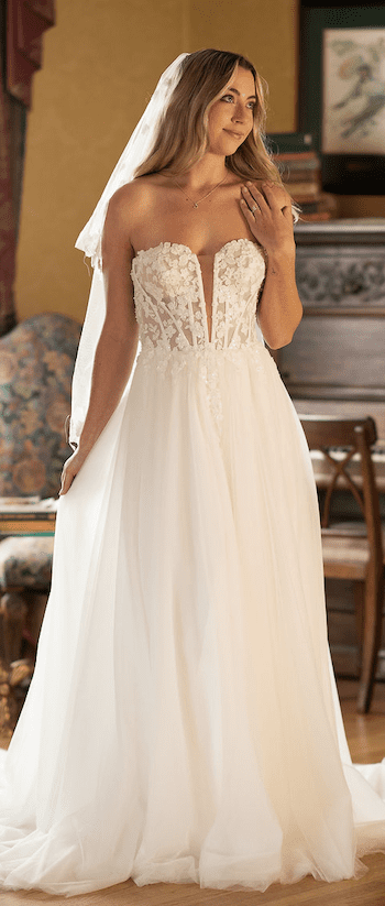 1738967100157-JoJo-EvelynBridal JoJo, an ivory A-line bridal gown by Sincerely Adorn, features a strapless sweetheart bodice with a deep plunge V-neck and illusion detailing. The bodice is adorned with delicate lace appliques and subtle beading, flowing into a soft, full tulle skirt with a chapel train. The model, with long blonde hair and a veil, poses elegantly indoors, looking to the side with a gentle smile.