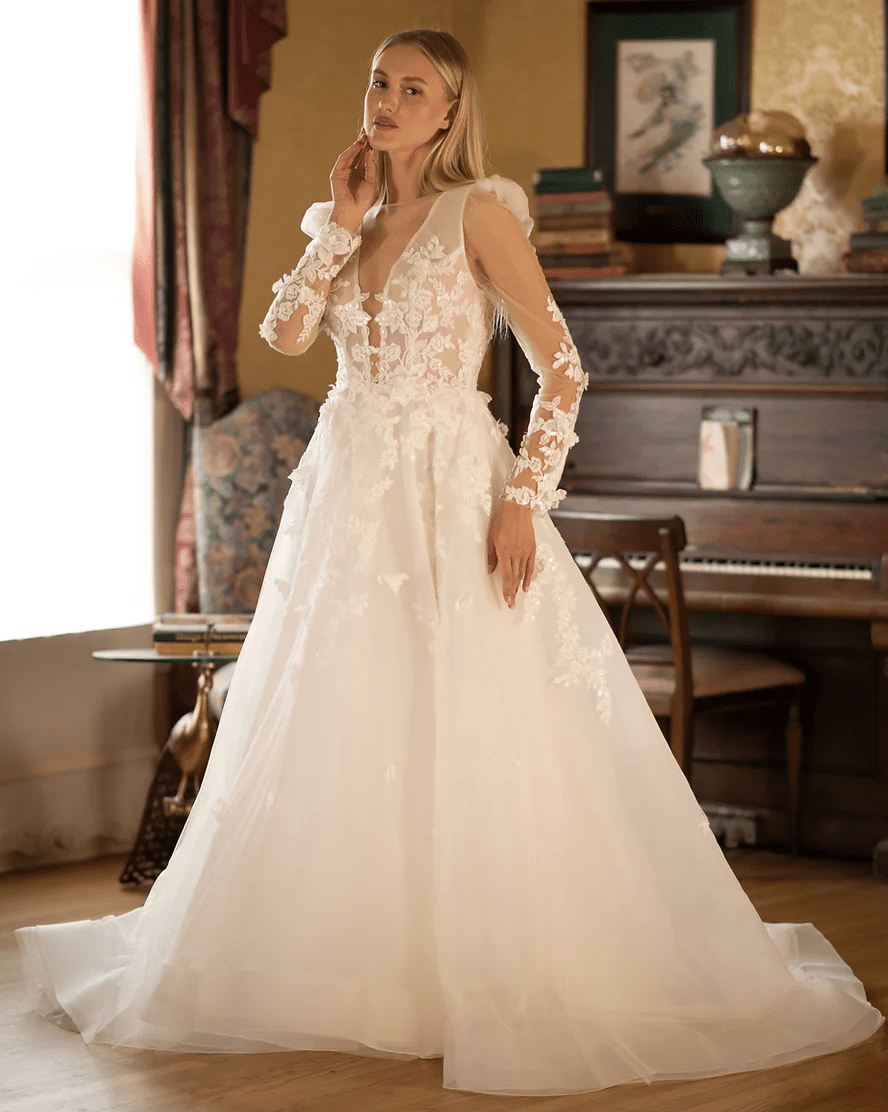 1743100306106-screen-shot-2025-03-27-at-2-31-18-pm Jolene, an ivory A-line bridal gown by Sincerely Adorn, features a plunging V-neckline over a sheer, illusion bodice adorned with delicate floral lace appliques, beading, and embroidery. The gown has sheer long sleeves with subtle ruffled shoulders and matching lace embellishments, while its full tulle skirt flows into a chapel-length train, showcasing scattered floral accents. The dress is modeled by a woman with long blonde hair, posing elegantly in a sunlit room.