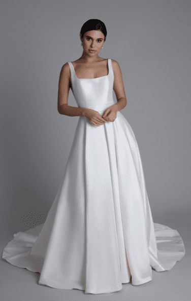 1743275550803-screen-shot-2025-03-29-at-3-10-21-pm Sincerity by Justin Alexander 44357A, an ivory Mikado ball gown wedding dress, features a square neckline, thick straps, a structured bodice, a low back with buttons, a full skirt with hidden pockets, and a chapel train. A model wears the dress, standing with hands clasped, against a plain grey studio background.