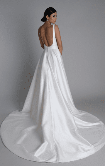 1743275550804-screen-shot-2025-03-29-at-3-10-26-pm 44357A, an ivory and nude Sincerity by Justin Alexander bridal ball gown, is seen from the back on a model looking over her right shoulder. This elegant stretch Mikado gown features a deep, low square back and a full line of Mikado-covered buttons gracefully extending down its chapel-length train. The design also incorporates flattering V-side cutouts and hidden pockets, perfectly blending modern style with classic romance and functionality.