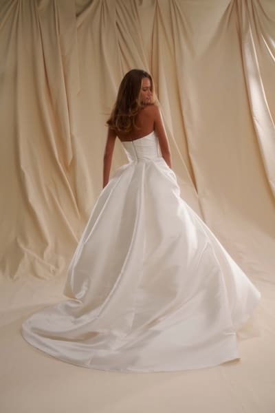 1747755381268-se007back SE007, a Diamond White Senstudio glamorous wedding gown made from lustrous Mikado fabric, featuring a strapless, hand-draped bodice, natural waist, and a voluminous ball gown skirt with a cathedral train. The dress is shown from the back on a model looking over her right shoulder, with the skirt flowing onto a cream floor against a draped fabric background.