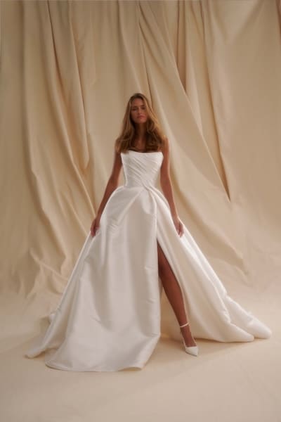 1747755381269-se007front Senstudio's SE007, a Diamond White Mikado bridal gown, featuring a strapless, ruched bodice, natural waist, a full ball gown skirt with a high slit revealing one leg, and a sweeping cathedral train, as worn by a model against a softly draped light-colored background.