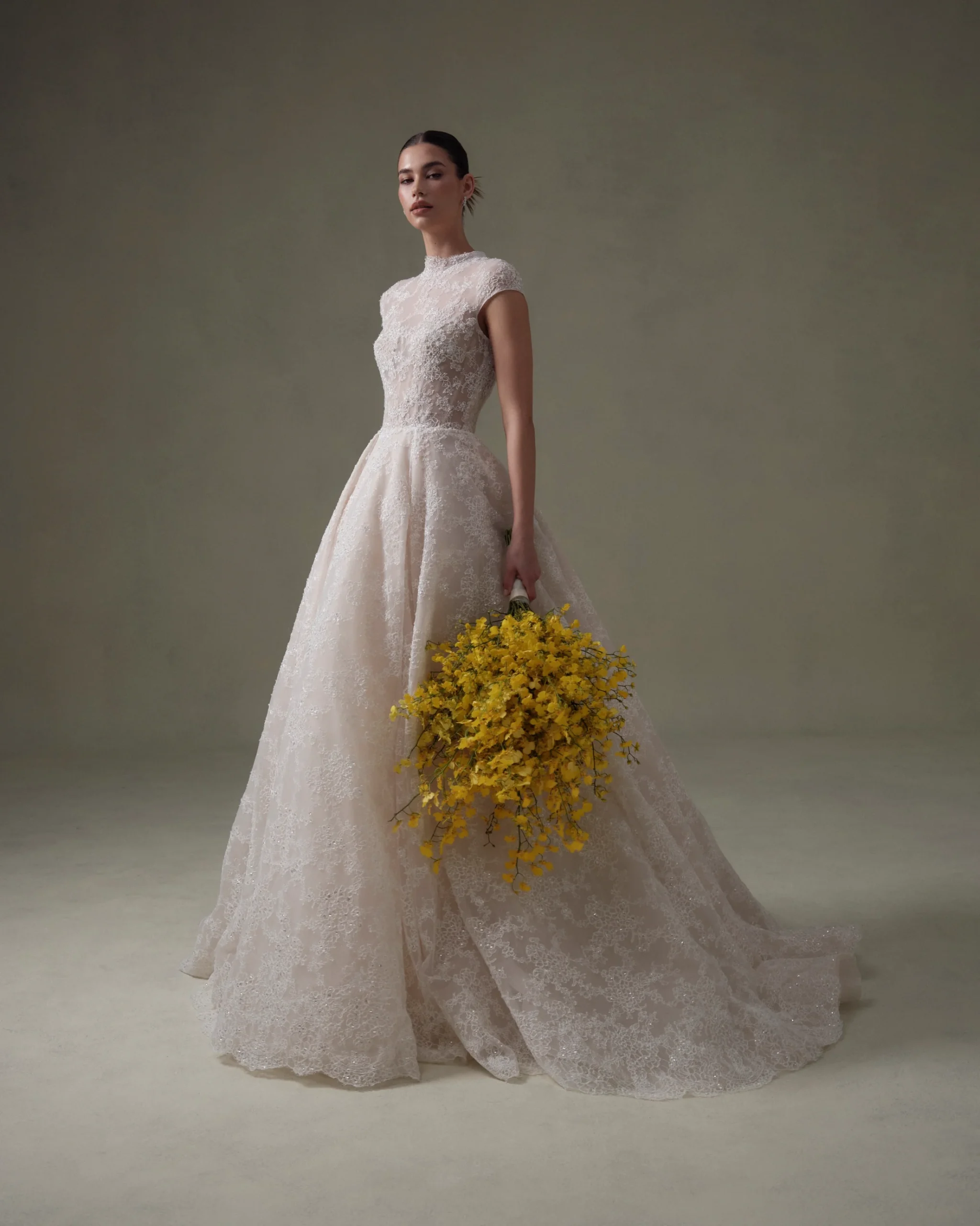 SE093 Privee, a Senstudio ivory and nude bridal ball gown, features a high neckline, elegant cap sleeves, and is adorned with intricate lace and sparkling beading from bodice to its chapel train. A model holds a vibrant yellow floral bouquet, looking directly at the viewer, against a muted green background.