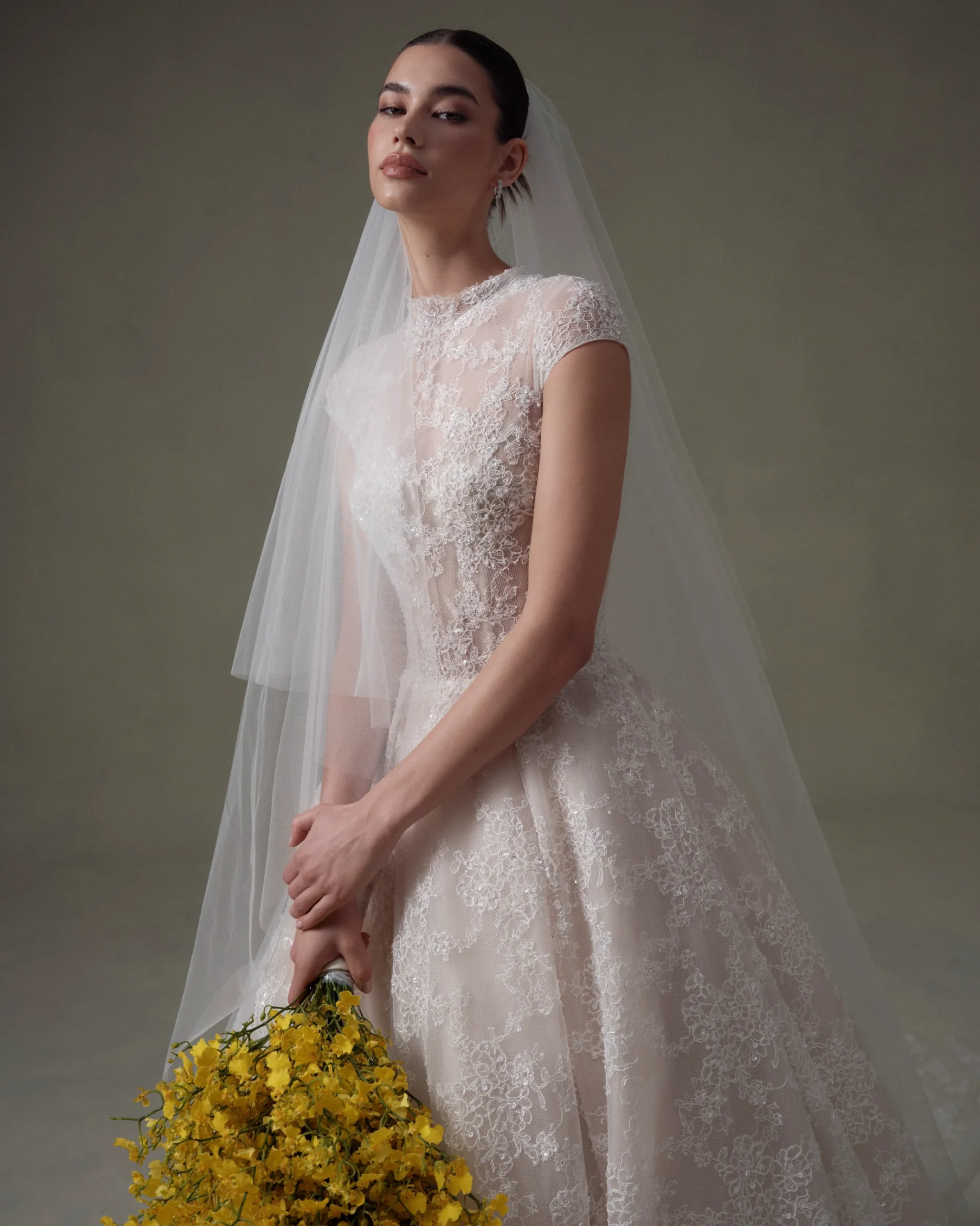 1756934444411-20250313NF4288 SE093 Privee, an Ivory/Nude ball gown wedding dress with a high neckline and delicate cap sleeves. The gown is adorned with intricate beaded lace throughout. A model with her hair in a sleek updo and natural makeup looks directly at the viewer, holding a vibrant yellow bouquet.