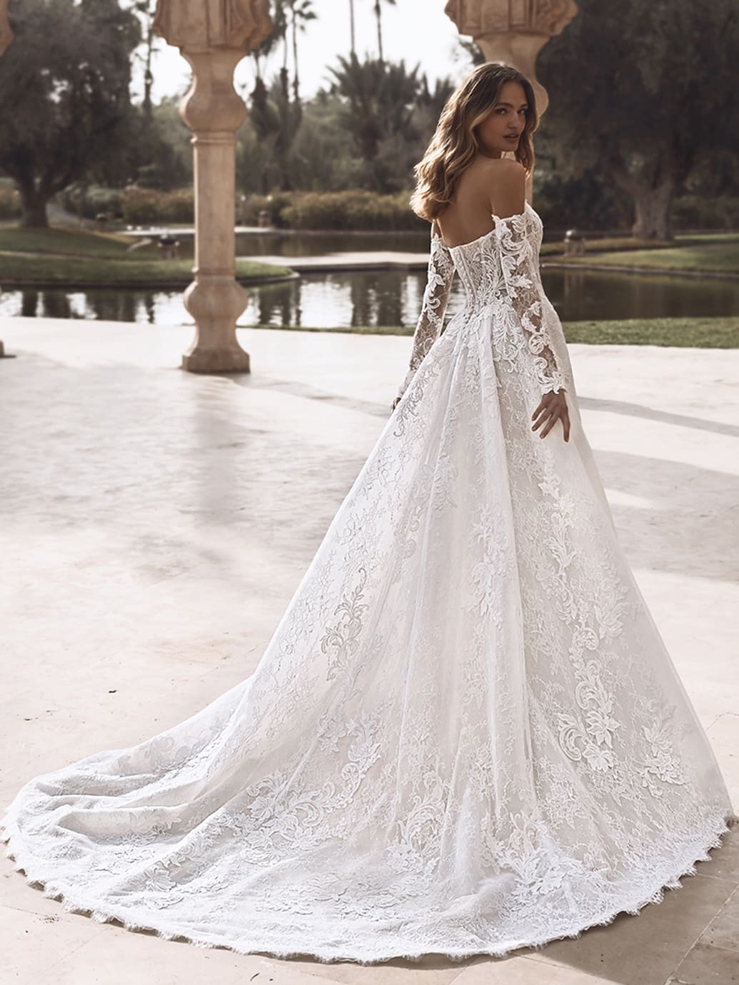 1758909380960-26_Enzoani_PRO_VANESSA_Bac_Web Vanessa, an ivory and sand/nude bridal gown by Enzoani, featuring an A-line silhouette crafted from delicate botanical lace and shimmering glitter tulle. The model wears the gown with its detachable off-the-shoulder long lace sleeves, turning to look over her shoulder, showcasing the dress's dramatic sculptural cathedral train that trails elegantly behind her on a paved outdoor area.