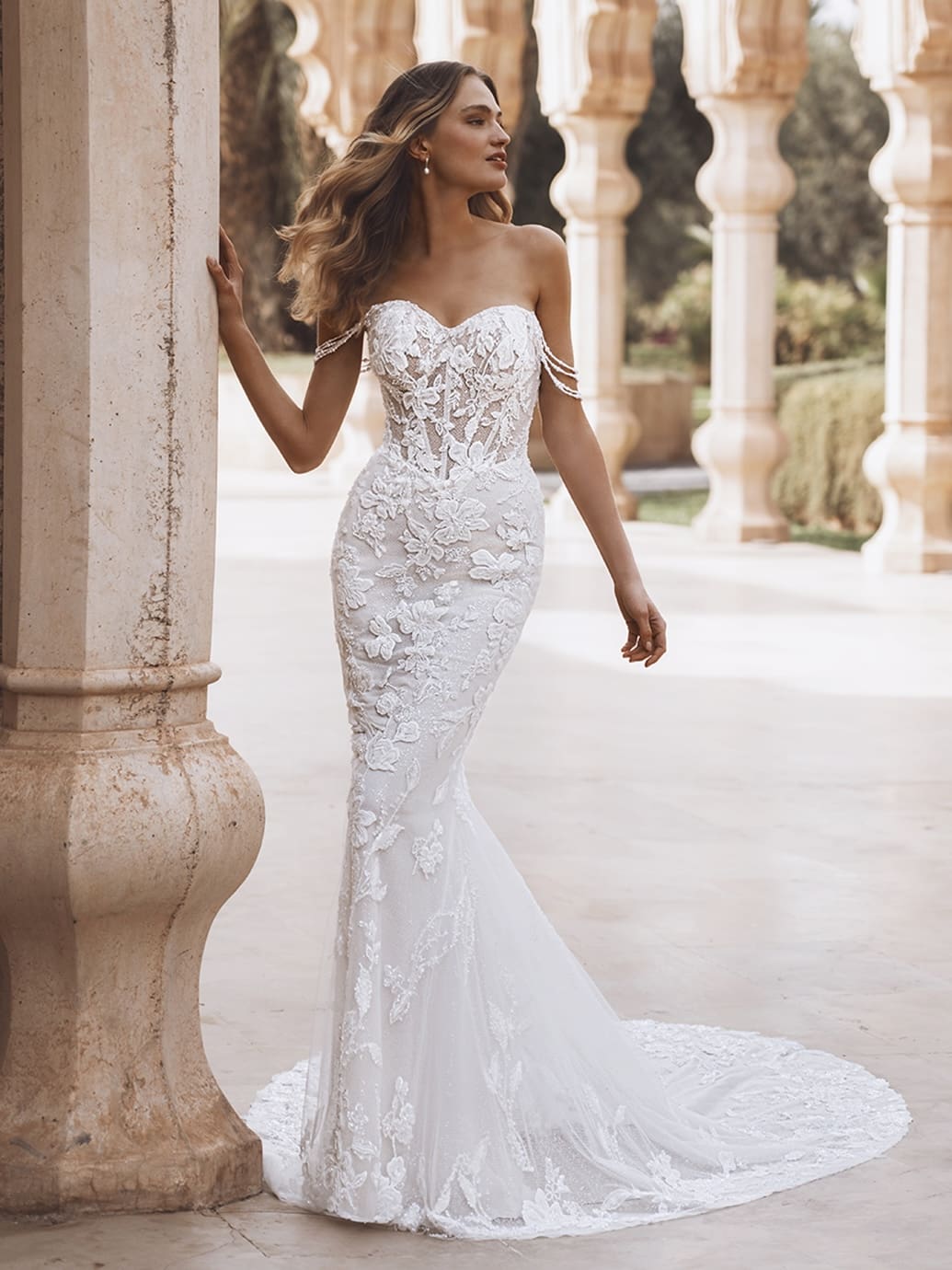 1758914733302-26_Enzoani_PRO_VIOLET_Fro_Web Violet, an ivory, sand, and nude fit-and-flare bridal gown, features a sweetheart neckline, an illusion corset bodice with intricate floral lace and embroidery, and delicate off-the-shoulder beaded straps. The gown's silhouette hugs the body before flaring into a chapel-length train adorned with matching lace. The model wears the dress outdoors, leaning against a stone column, with arched architectural details in the background.