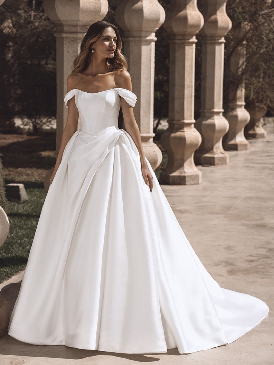 Viviana, an ivory Enzoani bridal ball gown crafted from luxurious Mikado fabric, featuring a strapless corset bodice with an off-the-shoulder scoop neckline and subtle ruching at the waist. The voluminous skirt flows into a dramatic cathedral train, detailed with buttons.
