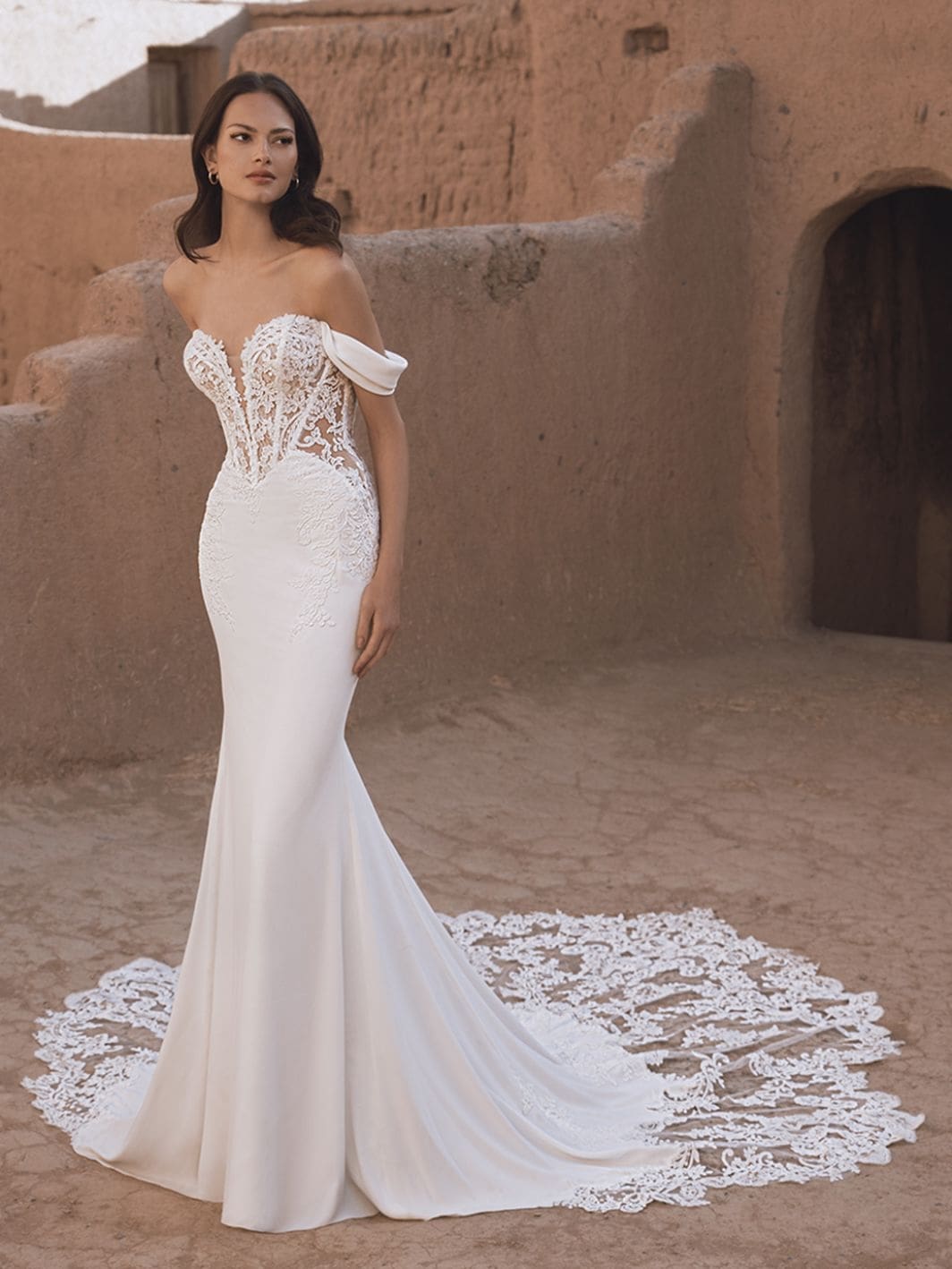 1758916009199-26_Blue_PRO_TAMMY_Fro_Web_1064_1418_80_s_c1 Tammy, an Ivory/Nude Fit n Flare wedding gown. It features a sweetheart plunge neckline with a sheer illusion lace bodice showcasing visible corset detailing, complemented by delicate off-the-shoulder draped straps. The smooth Georgette skirt flows into an elaborate train, intricately detailed with lace appliques. The gown is worn by a model with dark hair, standing gracefully outdoors against an ancient, earthy-toned architectural backdrop.