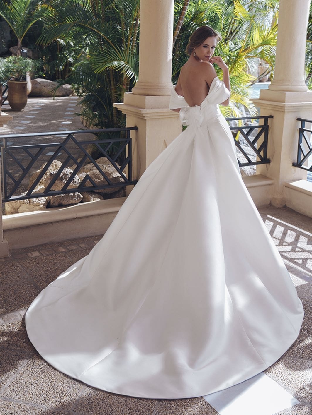 1758917666872-25-blue-pro-sevilla-sleeves-bac-web-1064-1418-80-s-c1 Sevilla, an ivory A-line wedding gown crafted in luxurious Mikado, features delicately draped, detachable off-the-shoulder straps, an open corset back with a tulle bow, and an elegant draped bodice. The pleated skirt boasts hidden pockets and extends into a voluminous cathedral train. A bride models the gown from behind, looking over her shoulder at the viewer, on a patio surrounded by tropical foliage and pillars.